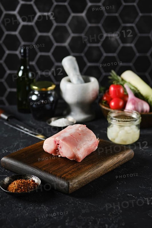 A raw rabbit saddle loin on a wooden cutting board in a kitchen setting. Fresh vegetables and spices surround it, providing a vibrant backdrop. The scene captures the essence of culinary preparation