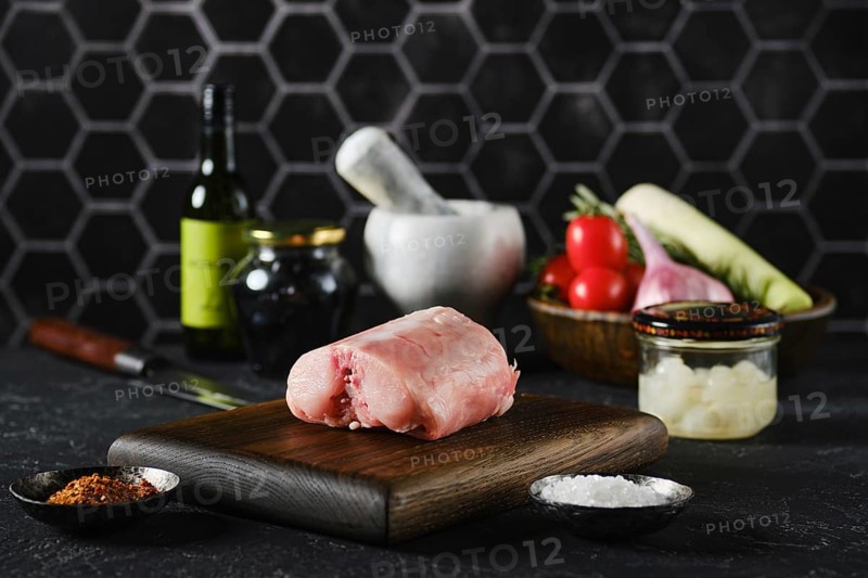 A fresh rabbit saddle on a wooden cutting board surrounded by fresh vegetables and cooking tools