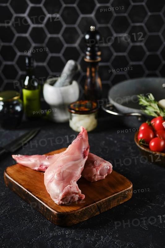 Raw rabbit hind legs on a wooden cutting board in a well-equipped kitchen. Fresh vegetables, oils, and various kitchen tools surround the meat, indicating preparation for a dish