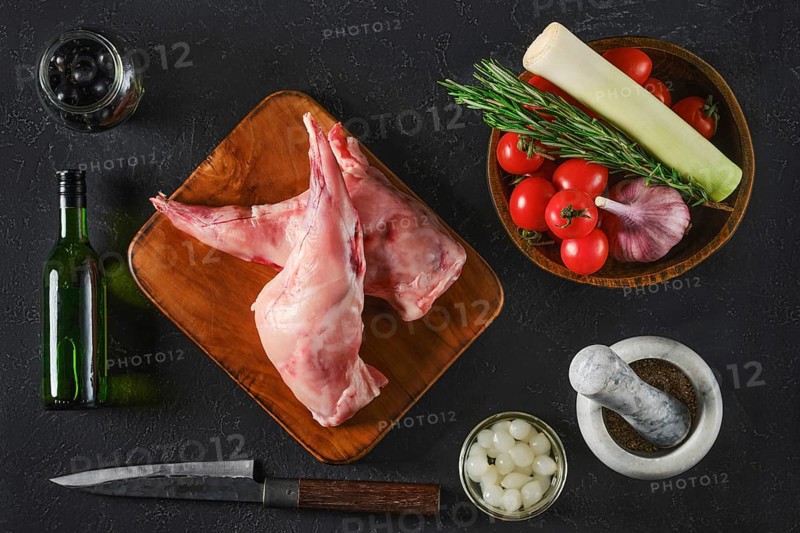 Top view of two raw rabbit hind legs are on a wooden board surrounded by fresh vegetables, herbs, and seasonings. The scene captures ingredients for a flavorful meal preparation on a dark surface