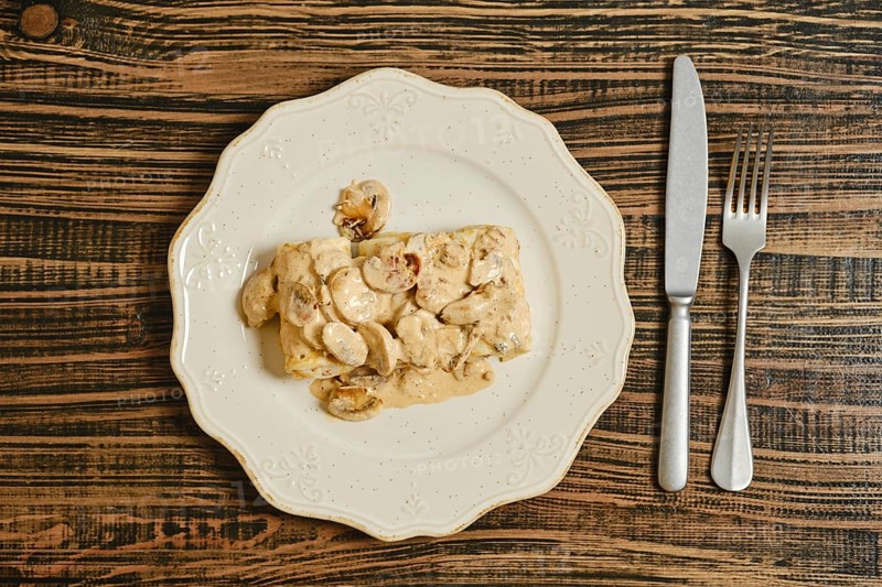 Crepes filled with seasoned meat, topped with a rich and creamy mushroom sauce. The dish is elegantly presented on a white plate against a wooden table