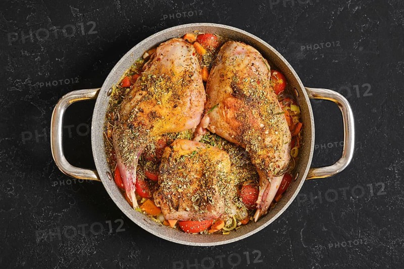 Two rabbit hind legs and a saddle are seasoned with herbs and spices, placed on a bed of colorful vegetables in a pan. The dish is ready to be cooked, showcasing vibrant ingredients and rich textures