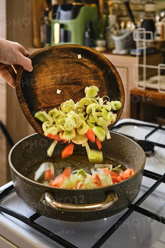 Sliced leeks and carrots are being poured from a wooden bowl into a pan on the stove. The warm light and homey setting create an inviting atmosphere for cooking a nutritious dish