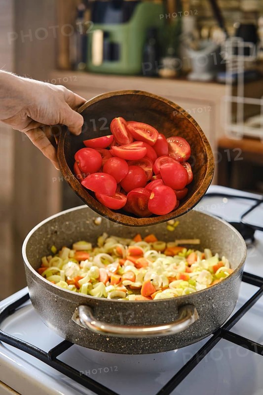 A person is adding chopped red tomatoes from a wooden bowl into a pot that is already filled with sauteed vegetables
