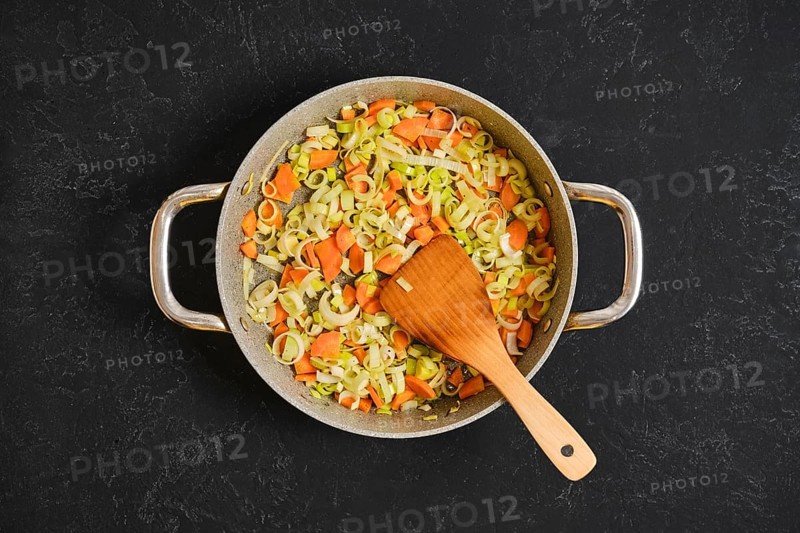 Chopped carrots and leeks are cooking in a large pan on a dark surface. A wooden spoon stirs the colorful vegetables