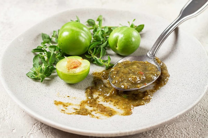 A spoon with tkemali sauce, on a plate, with cherry plum and mint, top view, no people