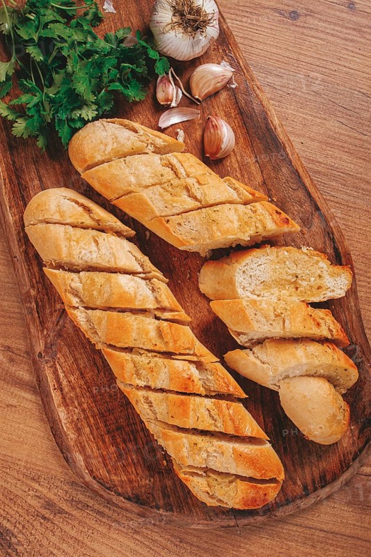 Garlic bread, baguette with garlic butter and herbs, on a wooden board, with herbs and garlic cloves