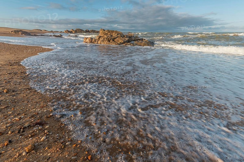 A wide sandy beach stretches out with scattered pebbles and beach shells. Gentle waves crash against the shore under a partially cloudy sky during late afternoon. Plage de l'Aubraie, Les Sables d'Olonne, Vendee, France