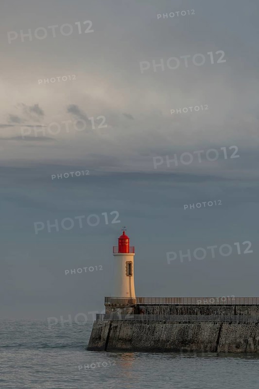 A lighthouse stands on a pier and casts its light over calm waters at dusk. The sky is filled with dramatic clouds, adding to the tranquil atmosphere of the coastal landscape. Les Sables d'Olonne, Vendee, France