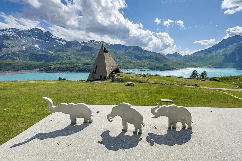 View from viewing platform to Mont-Cenis Pyramid Museum with stylised pyramid Museum in memory of Egypt campaigns of Emperor Napoleon Bonaparte and time in front of construction of reservoir, behind it Lac du Mont Cenis, in front on parapet small stylised figures of elephants Symbols for crossing of Alps Crossing of Alps by Hannibal, Mont Cenis Massif, Massif du Mont-Cenis, Graian Alps, Lanslebourg, Rhone-Alpes, France