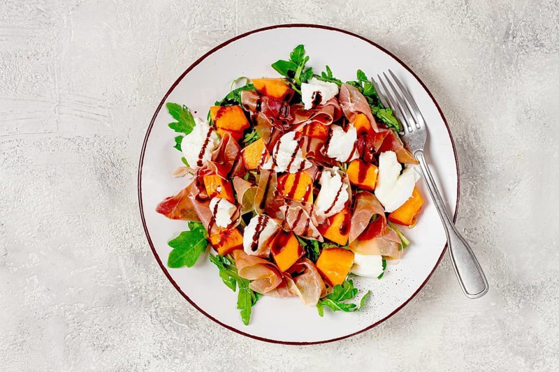 Salad with slices of ripe melon, prosciutto, mozzarella and fresh arugula, decorated with balsamic glaze