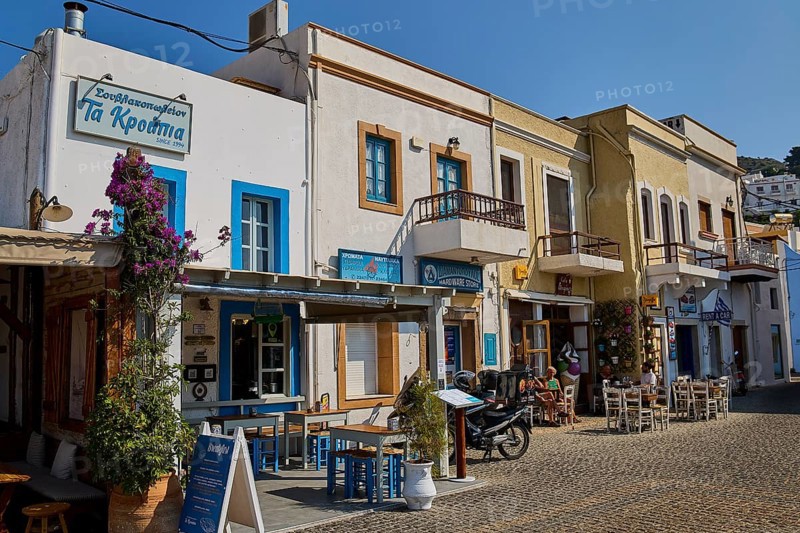 Row of historic buildings with restaurants and flowers on cobblestone street in sunshine, Agia Marina, Leros, Dodecanese, Greek Islands, Greece