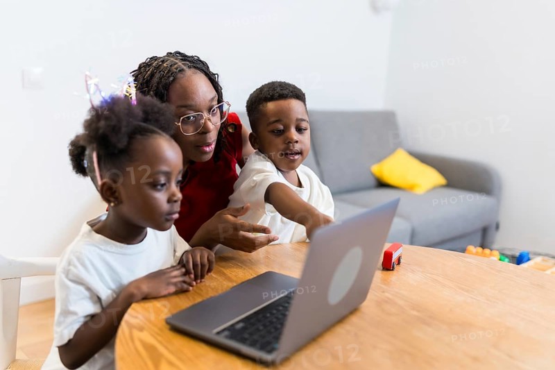 Young mother guiding her two children while using a laptop on a wooden table at home, fostering learning and connection in a cozy setting