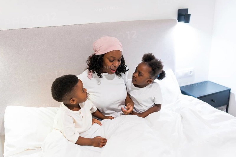 Mother undergoing cancer treatment, wearing a headscarf, sharing a heartwarming moment with her two children in bed, highlighting family support during challenging times