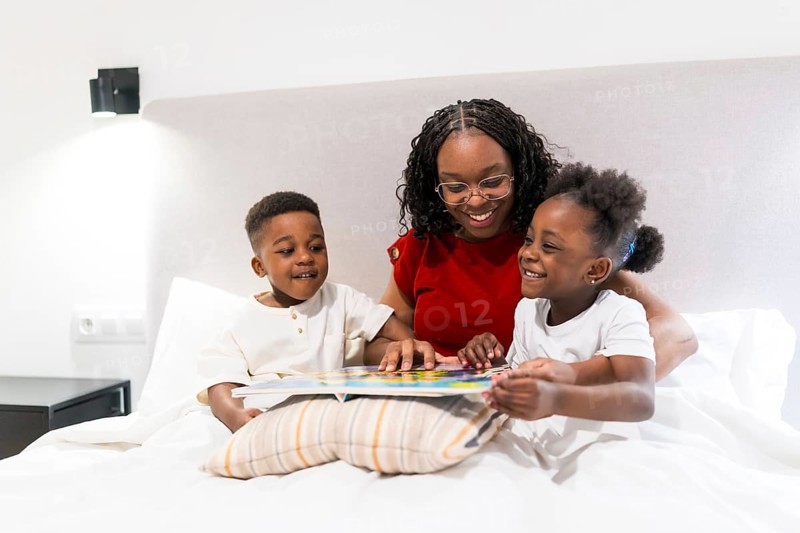 Smiling mother sharing a bedtime story with her two joyful children while cozily nestled on the bed, fostering love and connection