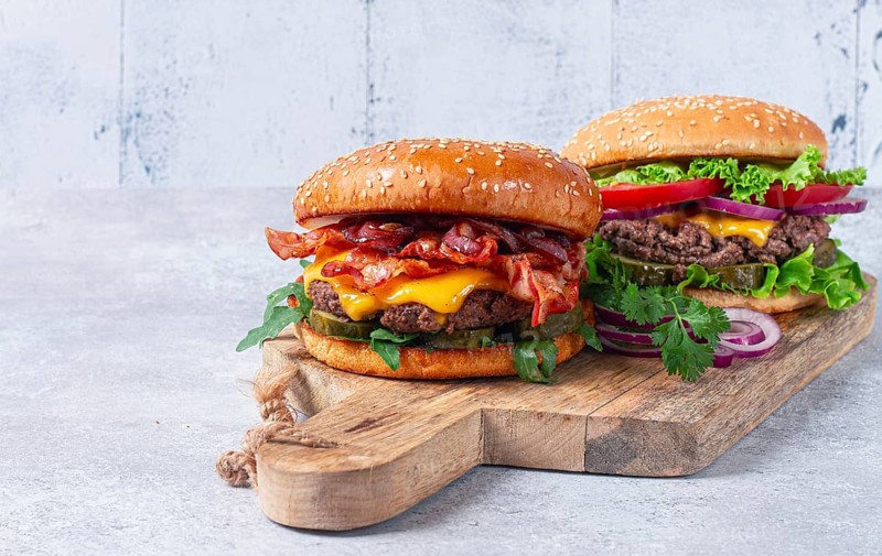 Two burgers with beef patty, fried bacon, cheddar cheese, tomatoes, on a light table