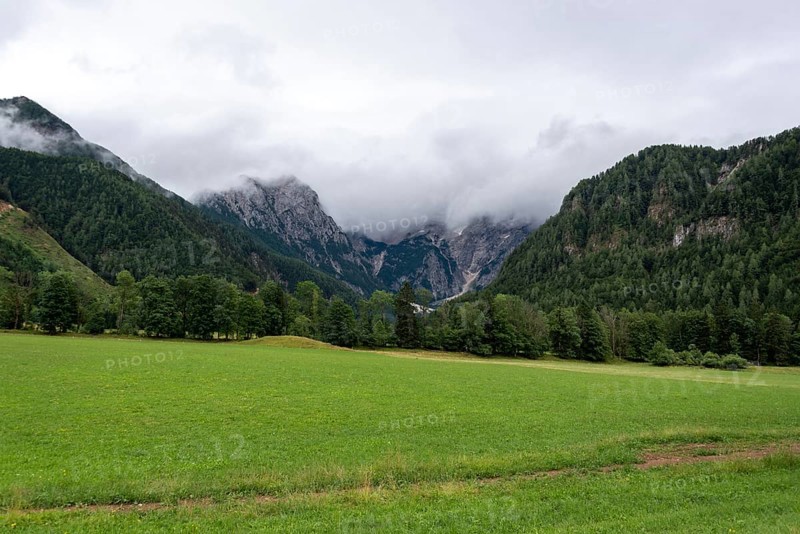 Green meadow against a mountain backdrop with clouds and wooded slopes, Zgornje Jezersko, Gorenjska, Slovenia