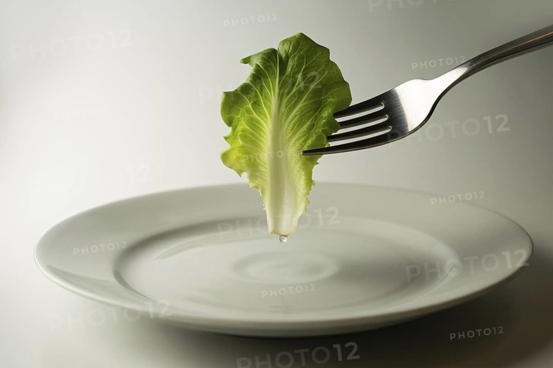 Close-up of fork holding lettuce leaf above white plate on white background. Concept of strict diet and unhealthy eating.. Generative ai, AI generated