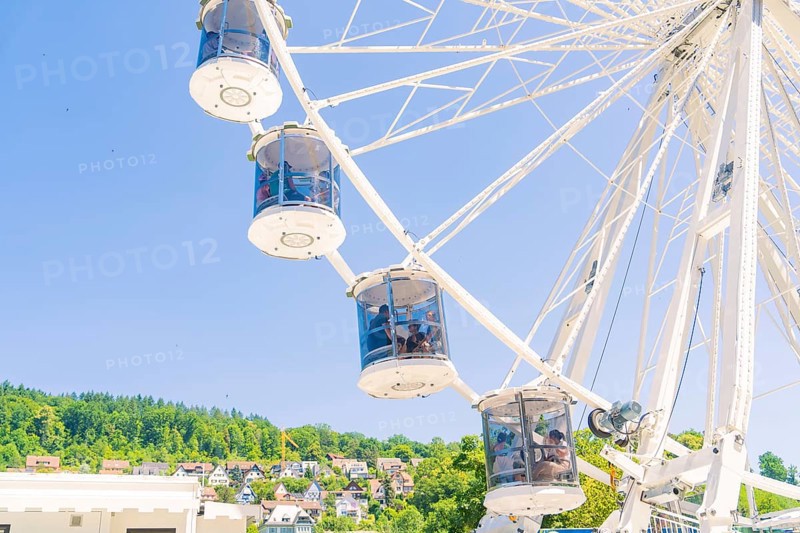 Ferris wheel with cabins in front of a blue sky, green trees and houses in the background, Calw, Germany