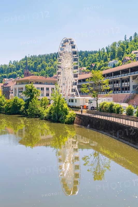 Ferris wheel reflected in the calm river, surrounded by buildings and wooded hills, Calw, Germany