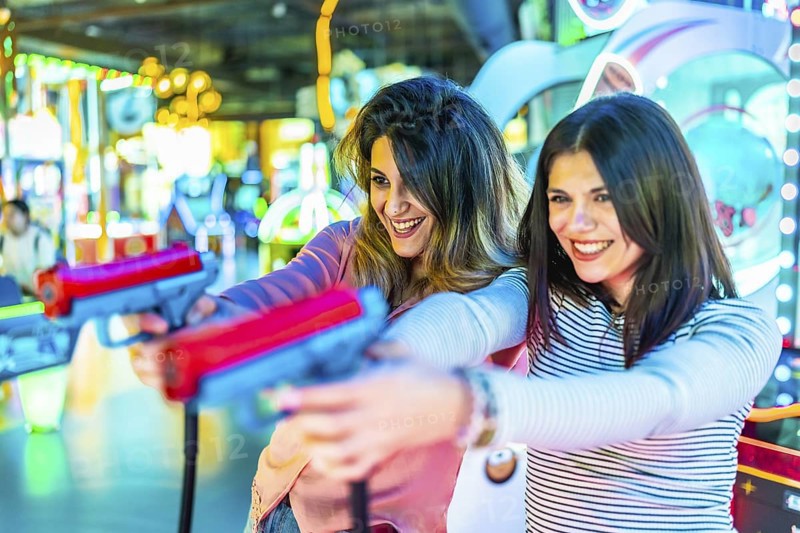 Two young women enjoying leisure time, playfully engaging with toy guns in a vibrant amusement arcade filled with colorful lights and laughter