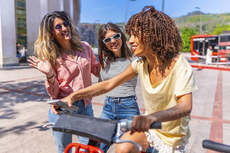 Three cheerful young women using smartphone and riding a sharing bike in a city center during summer holidays
