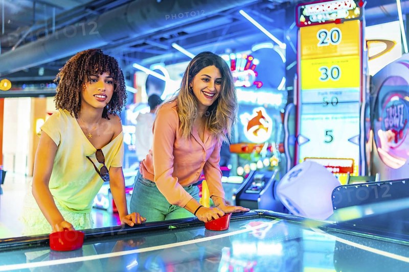 Two young women enjoying a friendly game of air hockey in a vibrant arcade, illuminated by colorful neon lights