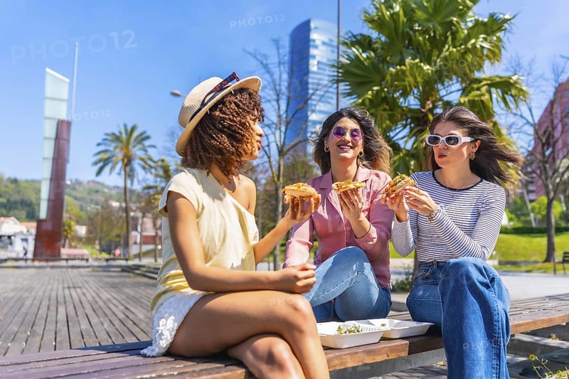 Three women savoring pizza slices while enjoying a sunny day on a park bench, sharing laughter and creating lasting memories in the city