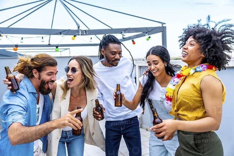 Cheerful multi ethnic friends laughing and enjoying drinks while celebrating together at a lively rooftop party under the summer sky
