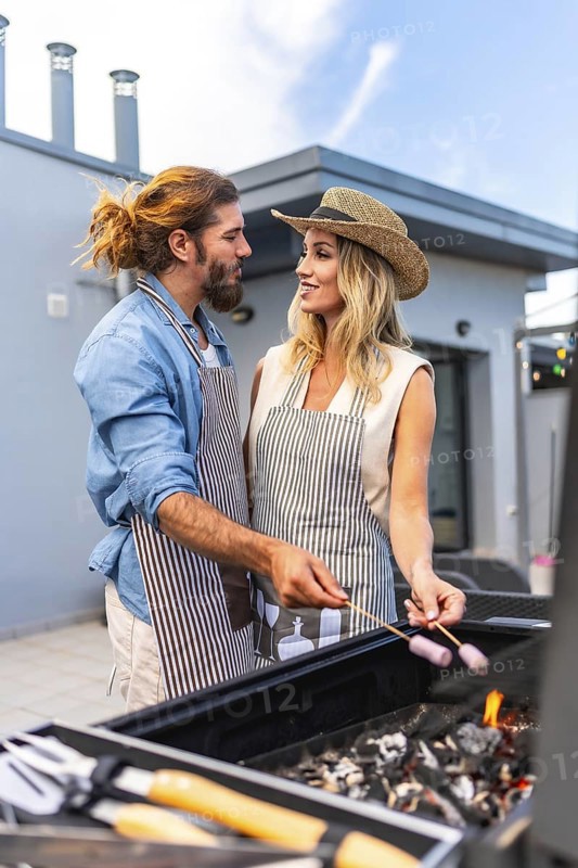 Happy couple enjoying a sunny summer day, grilling marshmallows on a rooftop barbecue, sharing laughter and creating joyful memories together