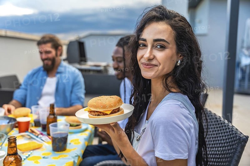 Young woman savoring a delicious burger while celebrating with friends at a lively rooftop barbecue party, enjoying the summer vibes