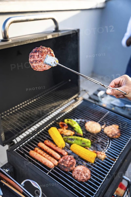 Hand using tongs to grill hamburgers, sausages, corn and peppers on a barbecue grill during a summer party