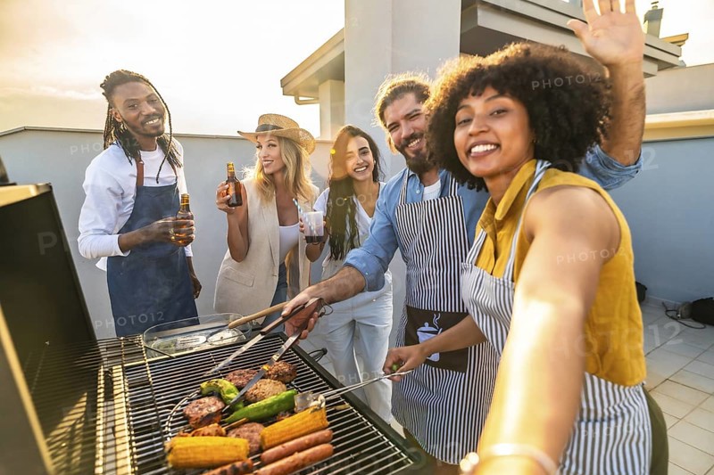 Cheerful multi ethnic friends are grilling meat and vegetables, drinking beer and taking a selfie on a rooftop at sunset