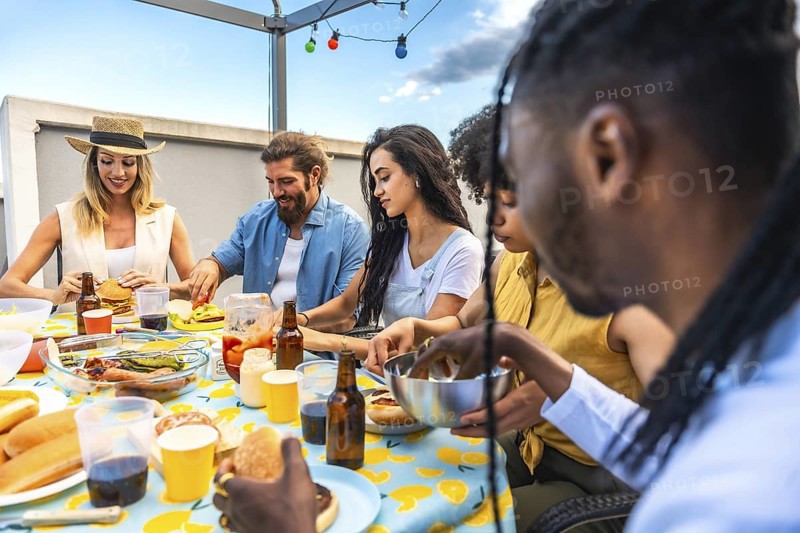 Multi ethnic friends enjoying a lively barbecue party on an urban rooftop, savoring delicious burgers and hot dogs while laughing together