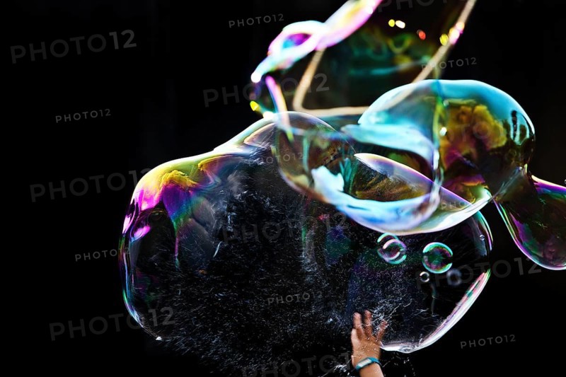 A hand bursts a large iridescent soap bubble against a black background, Recklinghausen, Germany