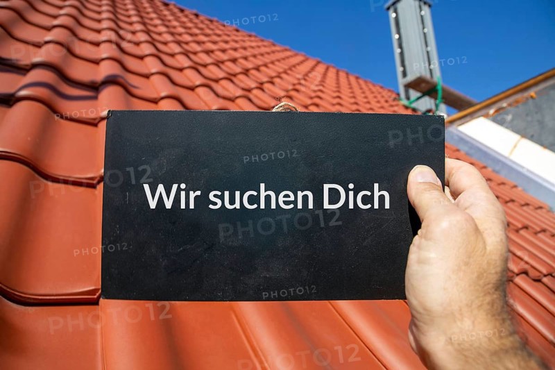 Employee search roofer: Man holding a sign (with added text) WE ARE LOOKING FOR YOU in front of a new tiled roof