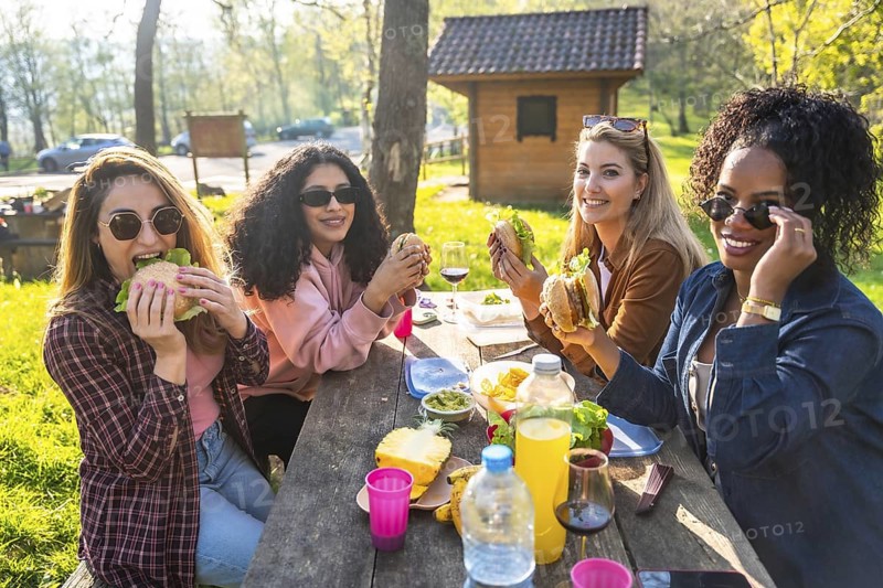 Four young women enjoying a picnic lunch outdoors, eating hamburgers and sharing good times together