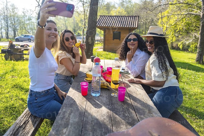 Four cheerful female friends enjoying a picnic outdoors, capturing the moment with a selfie