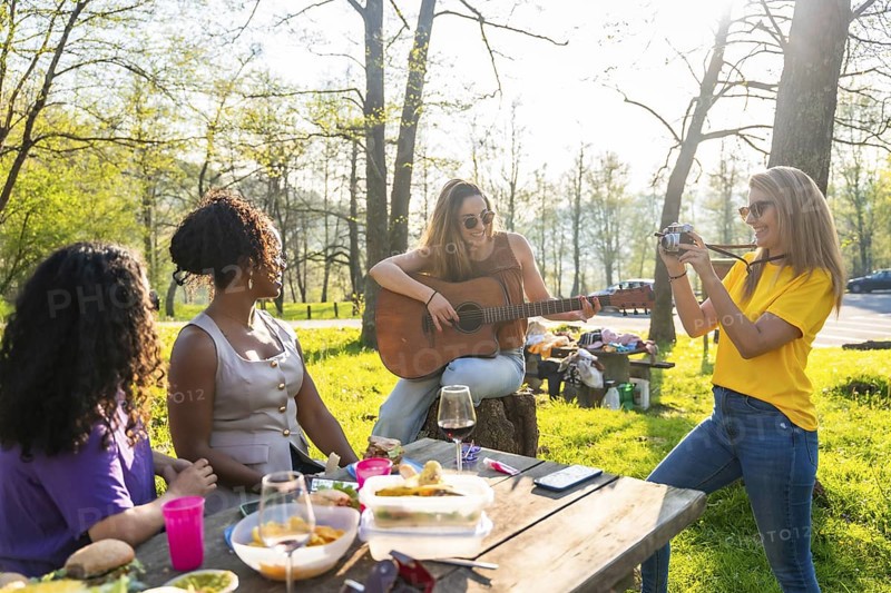 Four female friends enjoying a picnic in a park, with one playing guitar, another taking photos, and the others relaxing at the table