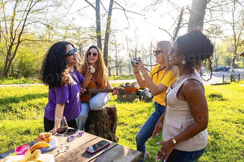 Four cheerful female friends are having a picnic in a sunny park, playing guitar, taking photos, and enjoying each other's company