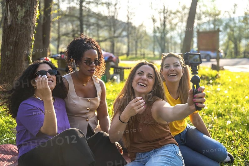 Four multi ethnic female friends are sitting on grass in a park, taking a selfie with an action camera mounted on a gimbal