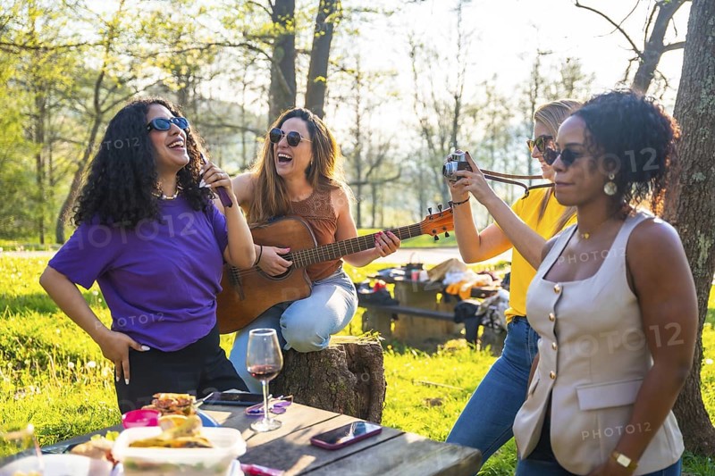 Four female friends enjoying a picnic in a park, singing, playing guitar, taking photos, and having fun