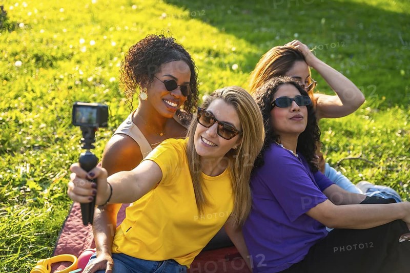 Four cheerful female friends are recording a video blog in the park using an action camera, enjoying their time together on a sunny day