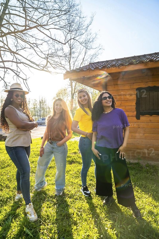 Four diverse female friends enjoying a sunny day together, posing confidently in front of a charming wooden house in a park