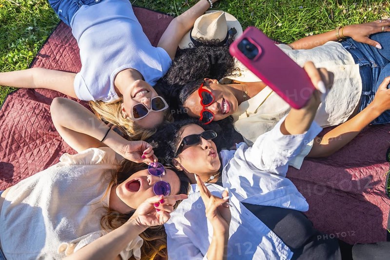 Four cheerful female friends are lying on a blanket in a park, taking a selfie with a smartphone and making funny faces