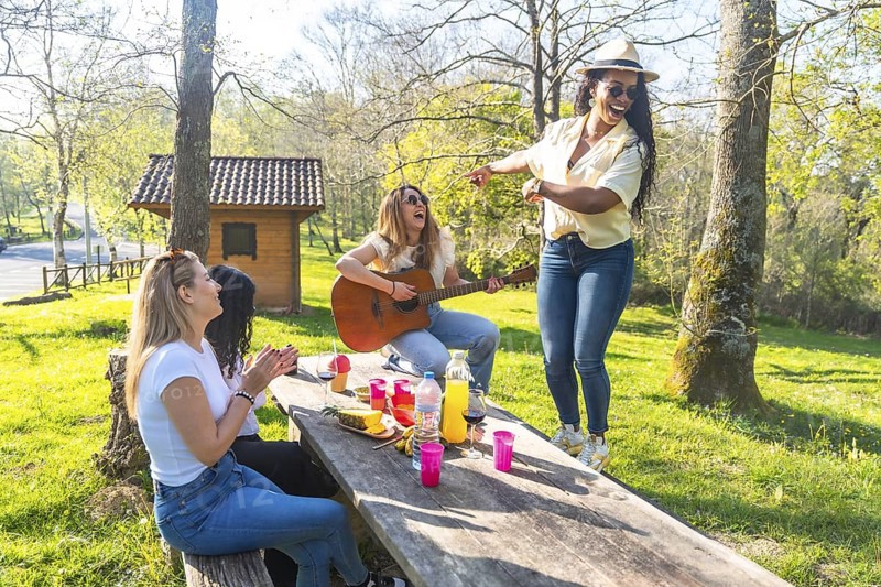 Four friends enjoying a picnic in a park, with one playing guitar, another dancing, and the others clapping and singing along