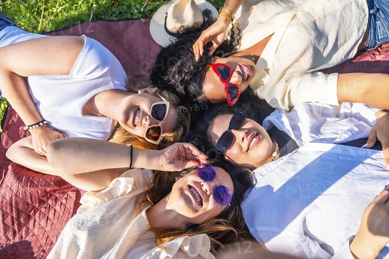 Four diverse young women lying together in a park on a blanket wearing sunglasses and smiling up at the camera