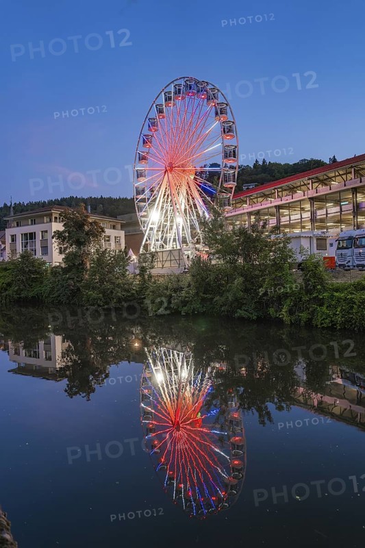 A pink illuminated Ferris wheel is reflected in the river, surrounded by municipal buildings, 950 years of Calw, Germany