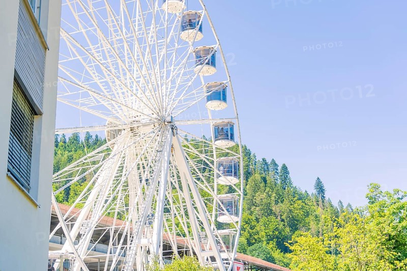 A large Ferris wheel stands next to a building with a wooded hill and clear sky in the background, 950 years of Calw, Germany