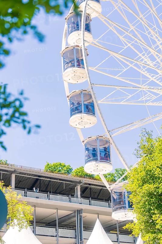 The Ferris wheel with glass cabins rises next to a large building surrounded by sunny trees, 950 years Calw, Germany